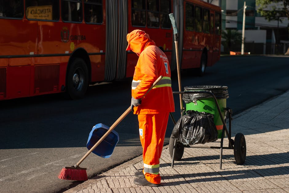 A street cleaning worker from Altındağ Belediyesi, dressed in an orange high-visibility vest with the company name printed on the back, is using a long-handled tool to sweep the pavement outside a h