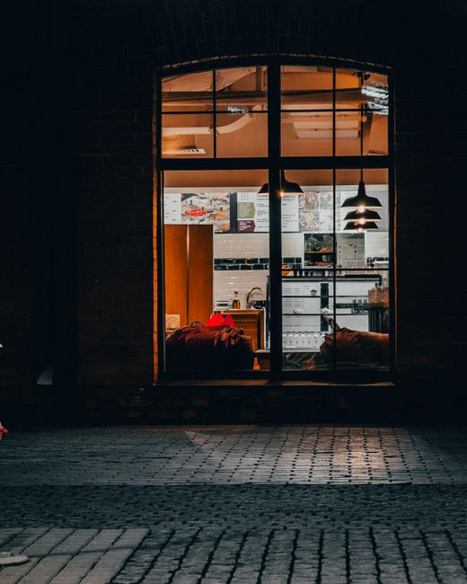 The image depicts a nighttime street scene outside Maltby Street Market, illuminated by warm, artificial lights from nearby shops and street lamps. A group of people are gathered on the sidewalk, with